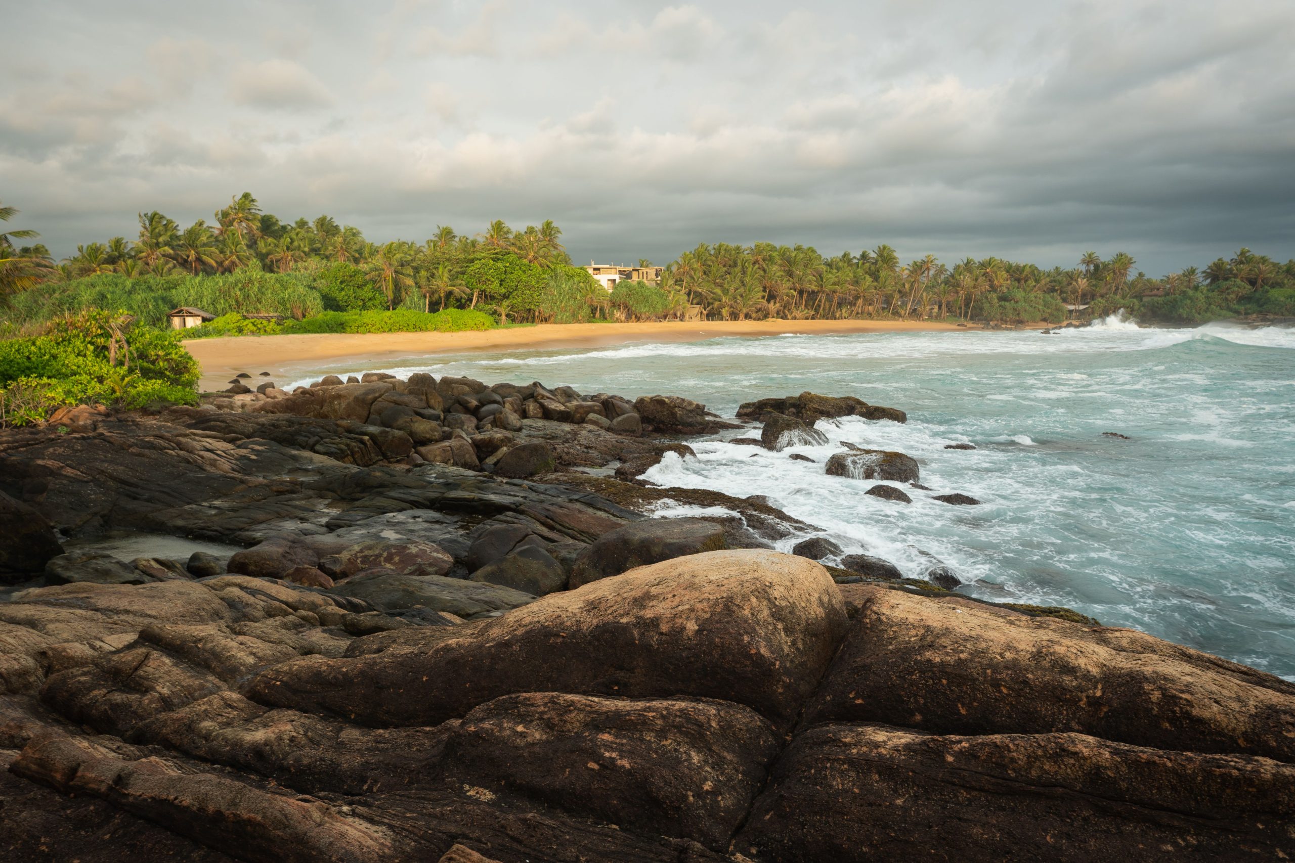 Explore a serene tropical beach in Sri Lanka with rocky shores and lush greenery under a dramatic sky.